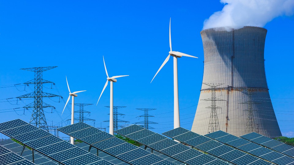 Solar panels with wind turbines in the foreground leading back to a atomic energy plant against a clear blue sky